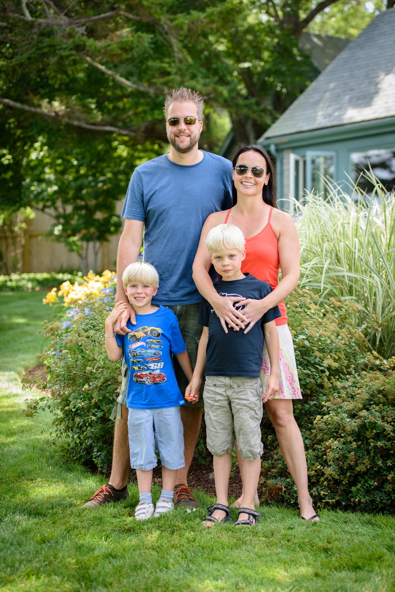 A family of four stands together in a garden, smiling at the camera.