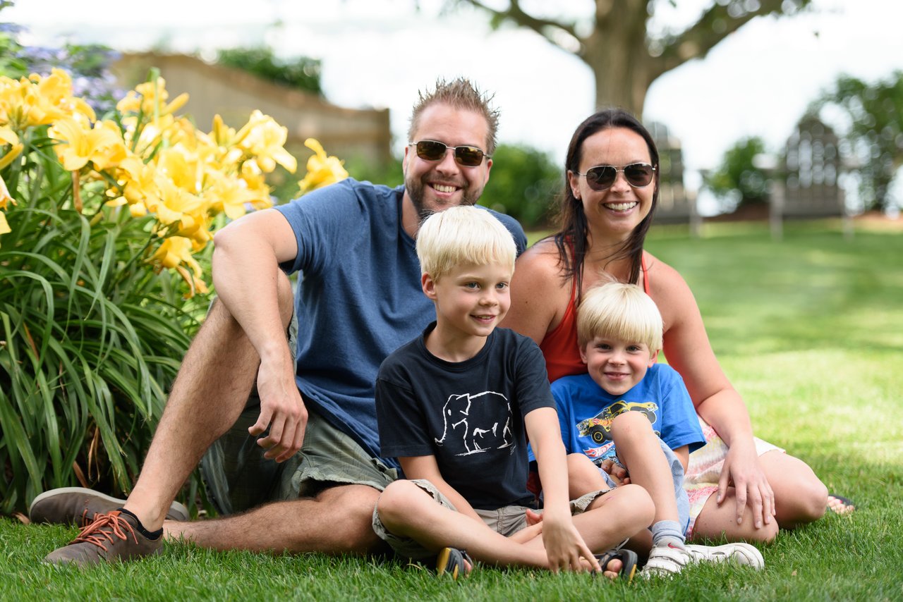 A smiling family of four sits on the grass, posing for a photo on a sunny day.
