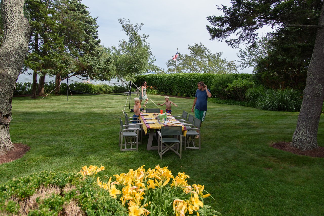 A family gathers in a backyard with a set dining table, children playing, and a man on the phone.