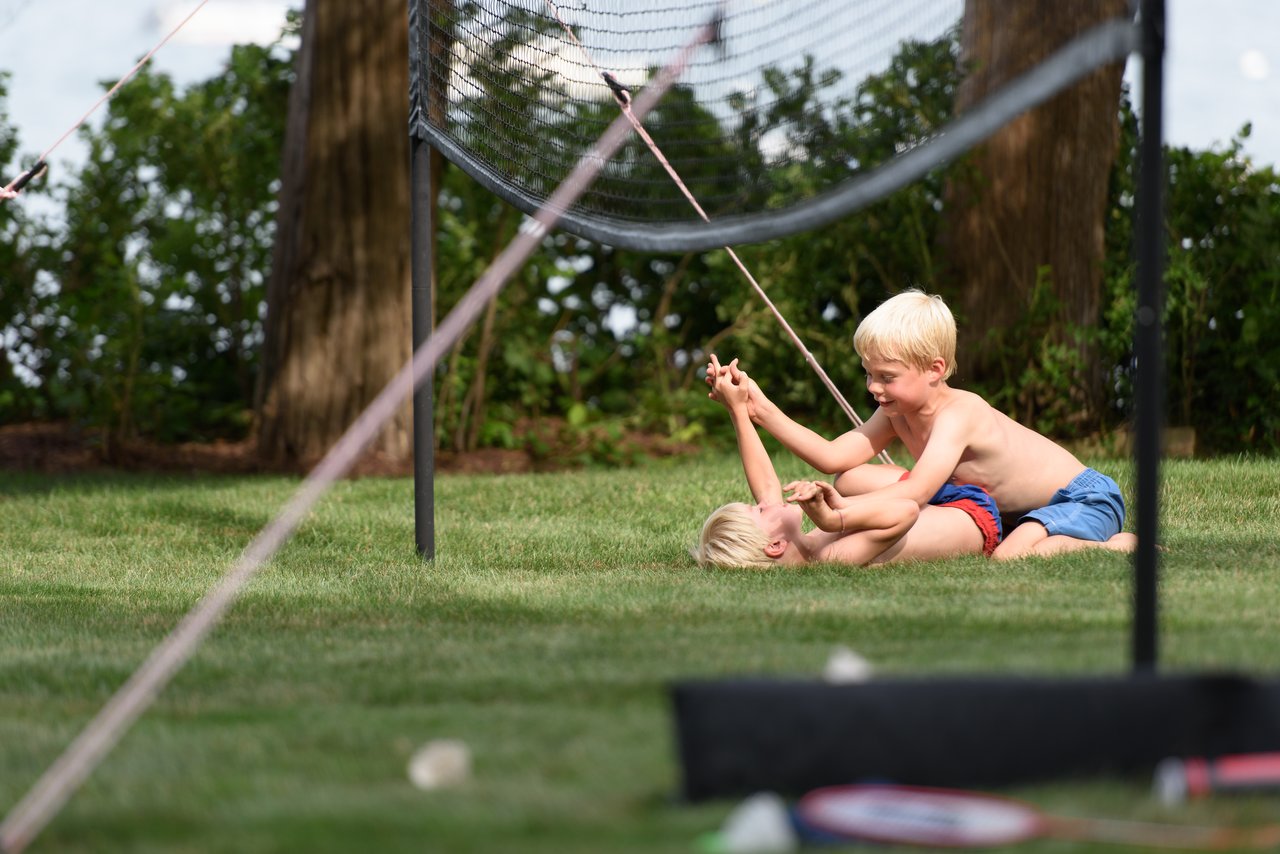 Two young boys in swim trunks playfully wrestle on the grass near a badminton net.