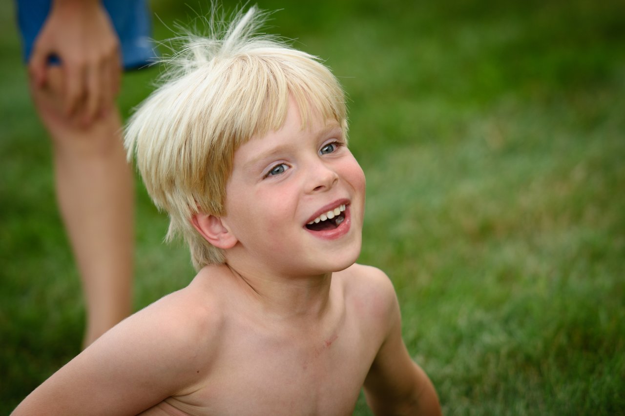A smiling, shirtless child with blond hair sits on the grass, looking up with excitement.