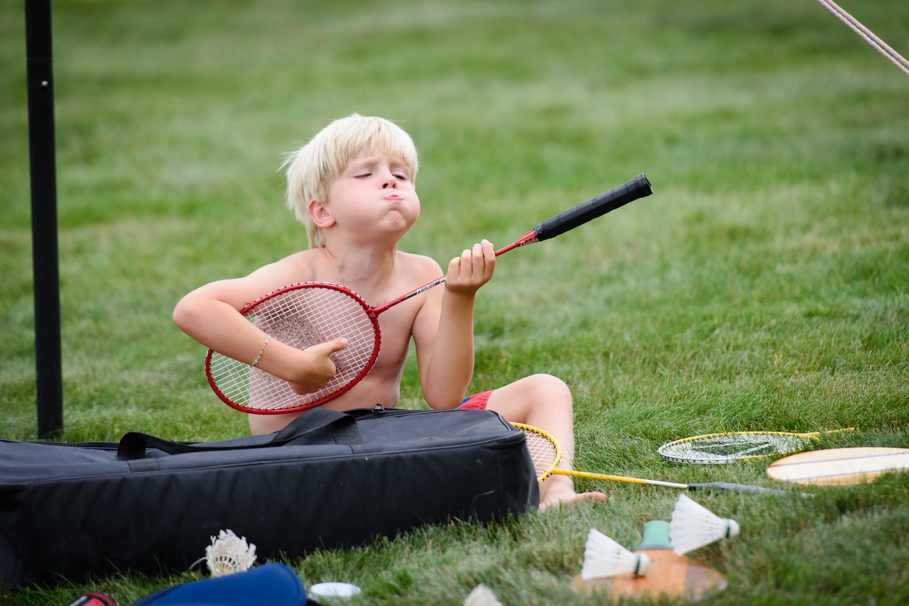 A shirtless child sits on the grass, holding a badminton racket and making a playful face.