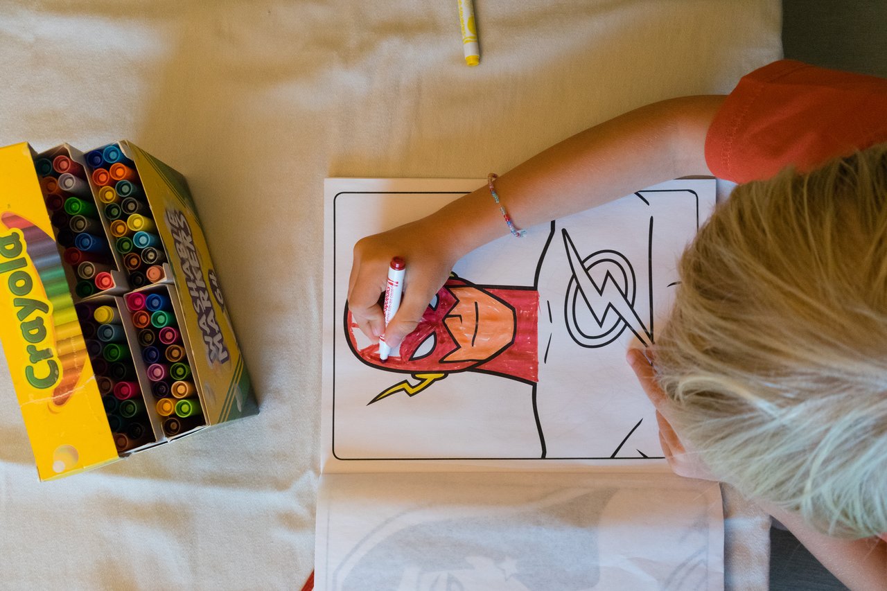 A child colors a picture of The Flash using a red marker, with a box of markers nearby.