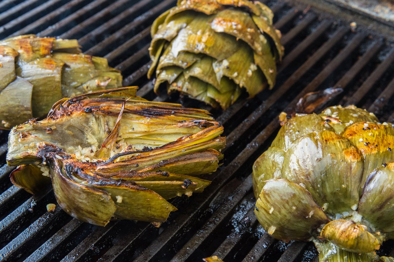 Grilled artichokes with seasoning on a barbecue grill, showing charred edges and a golden-brown surface.