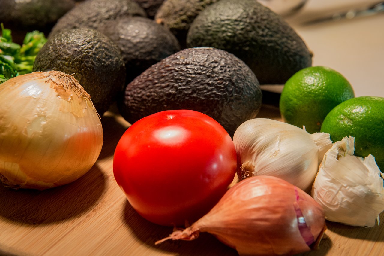 Fresh ingredients for guacamole, including avocados, tomato, onion, garlic, shallot, limes, and cilantro on a wooden surface.