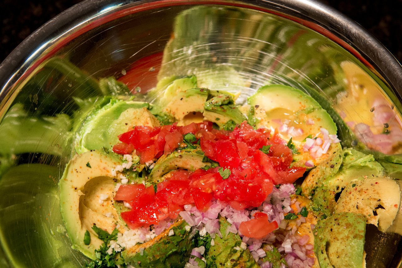 A bowl with avocado, chopped tomatoes, onions, and seasoning, ready to be mixed into guacamole.