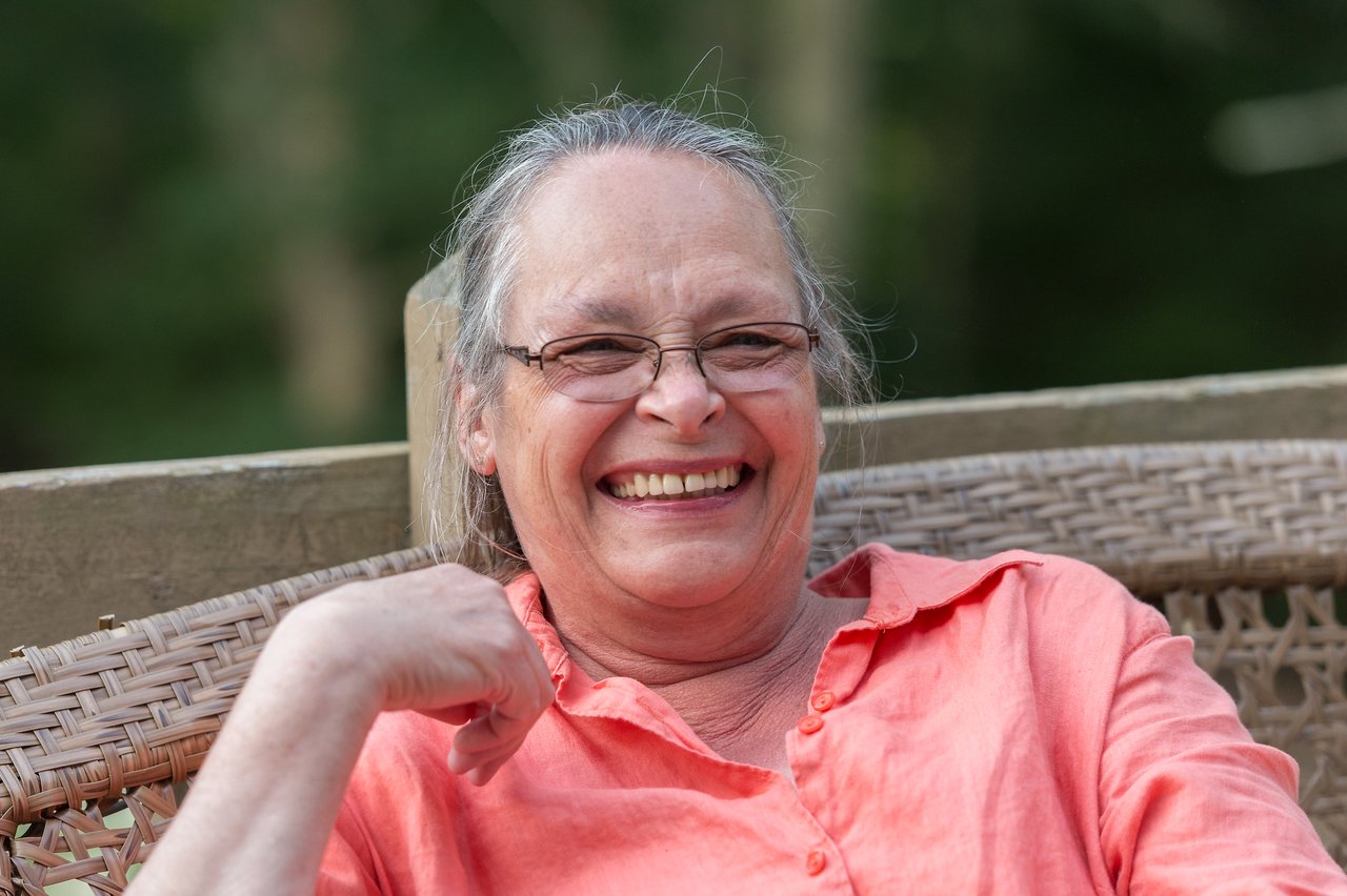 A woman wearing glasses and a coral shirt smiles while relaxing on a wicker chair outdoors.
