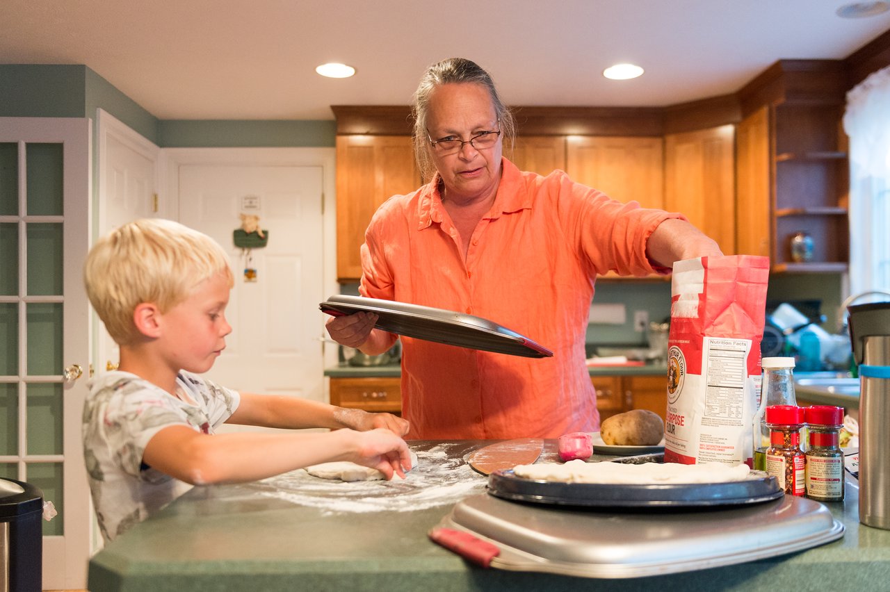 A woman and a child prepare pizza dough together in a kitchen, with flour and ingredients on the counter.