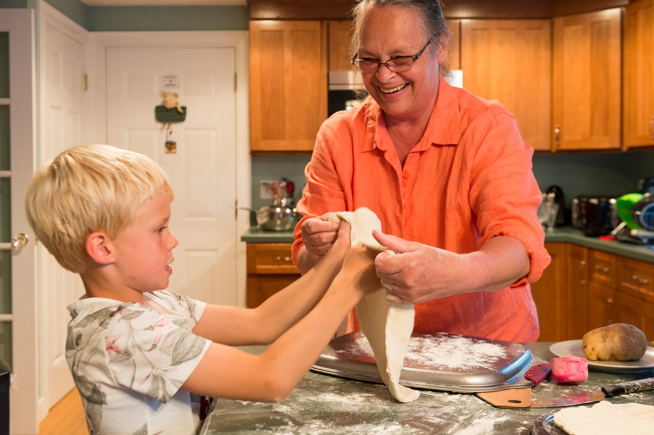 A smiling woman and a young boy stretch pizza dough together in a kitchen.