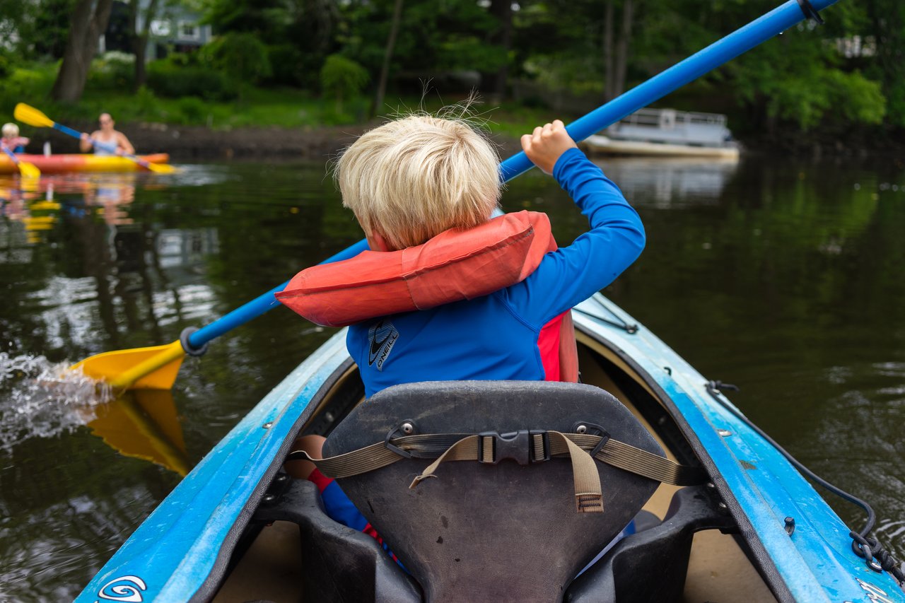 A child wearing a life jacket paddles a blue kayak on calm water, with other kayakers in the background.