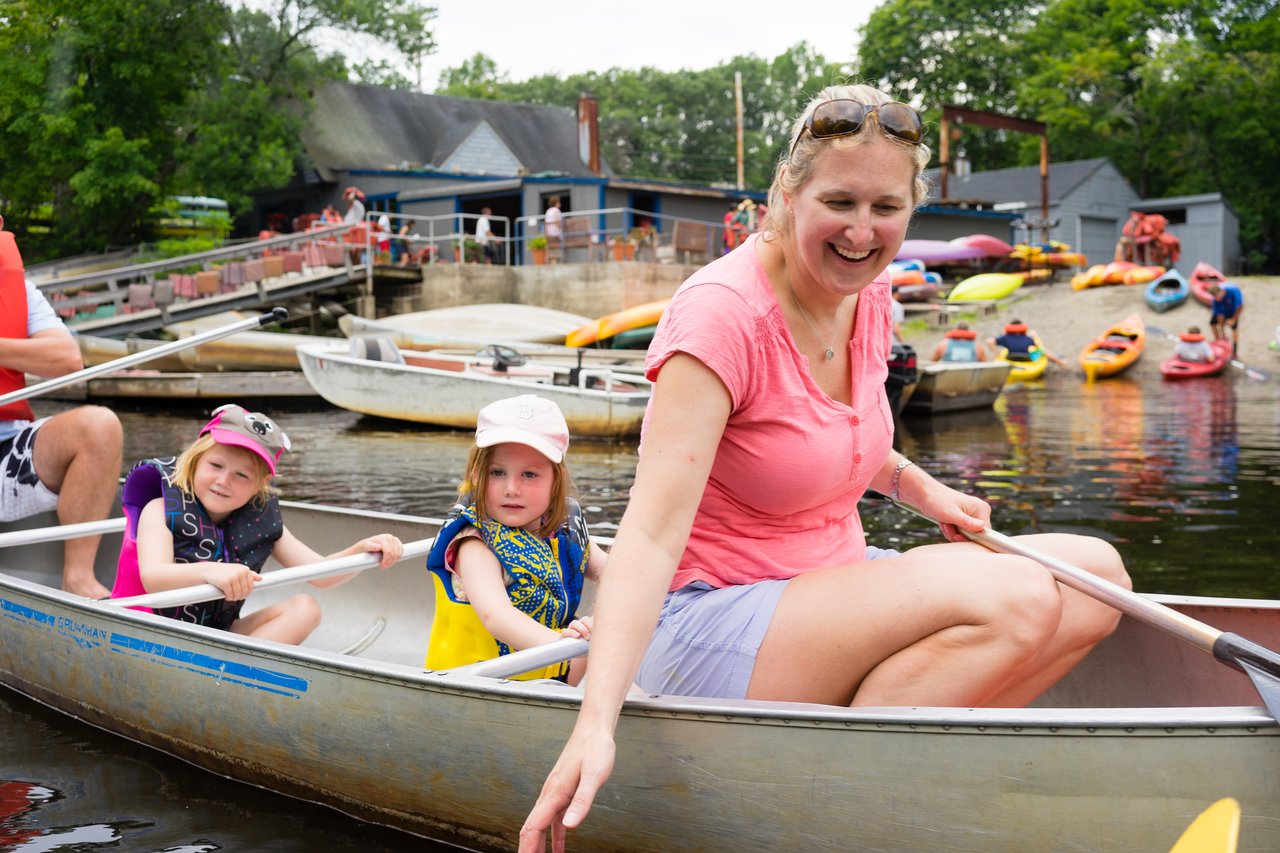 A smiling woman and two young children paddle a canoe on the water, surrounded by other boats and kayaks.