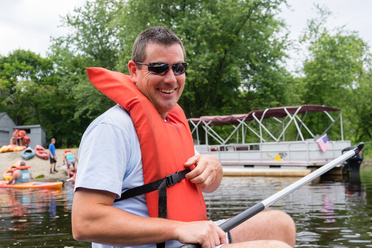A man wearing a life jacket and sunglasses smiles while holding a kayak paddle on the water.