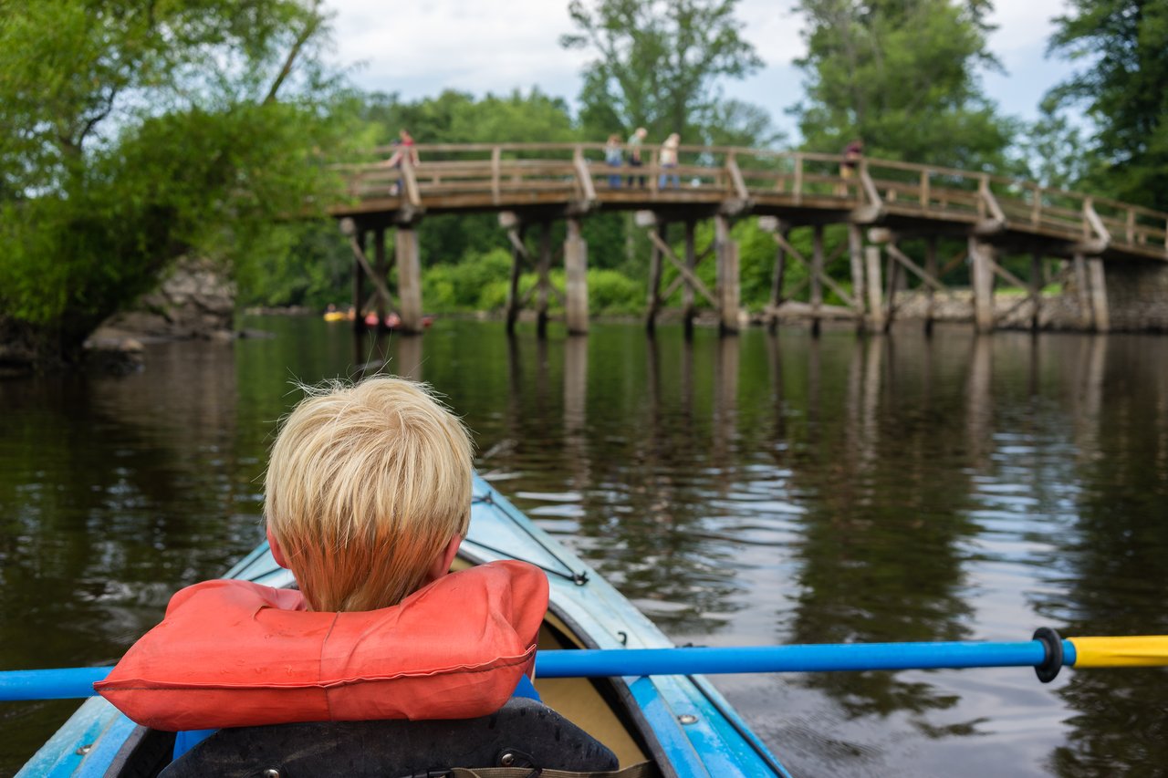 A person wearing a life jacket paddles a blue kayak on a river, approaching a wooden bridge.