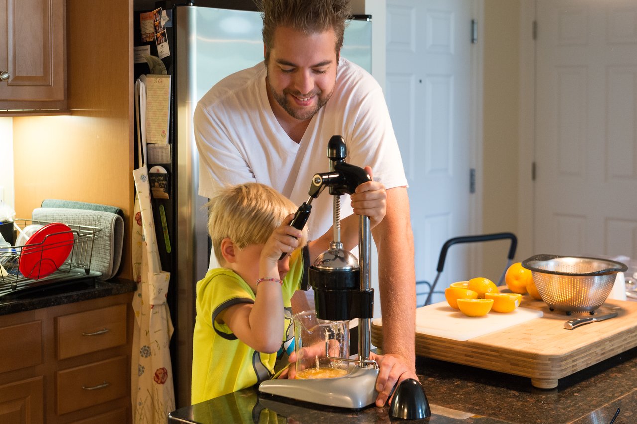 A man and a child use a manual juicer to squeeze fresh orange juice in a kitchen.