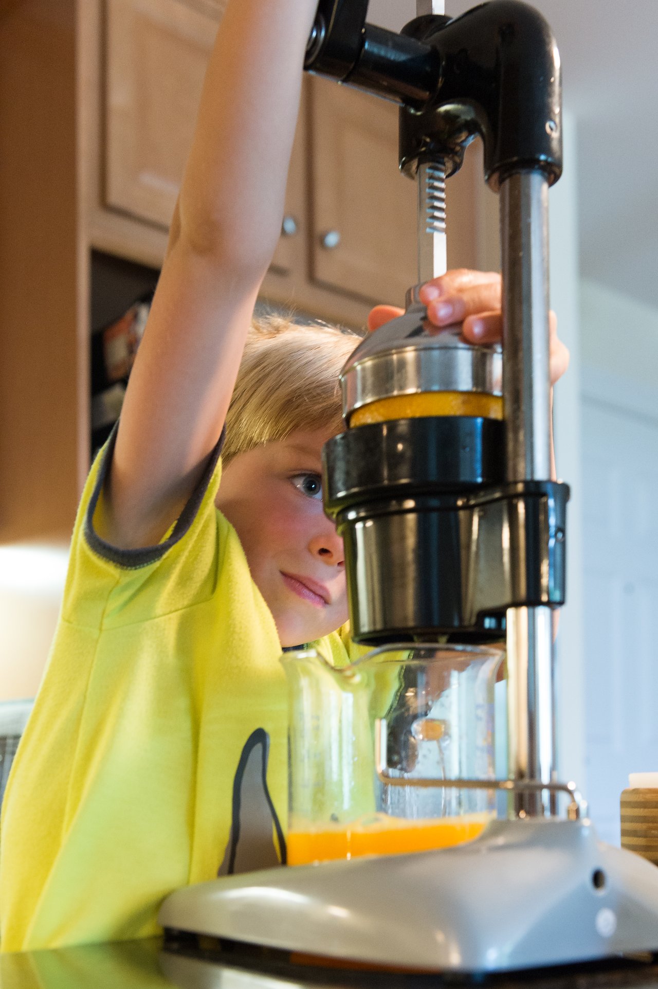 A child in a yellow shirt presses an orange juicer handle, squeezing fresh juice into a glass container.