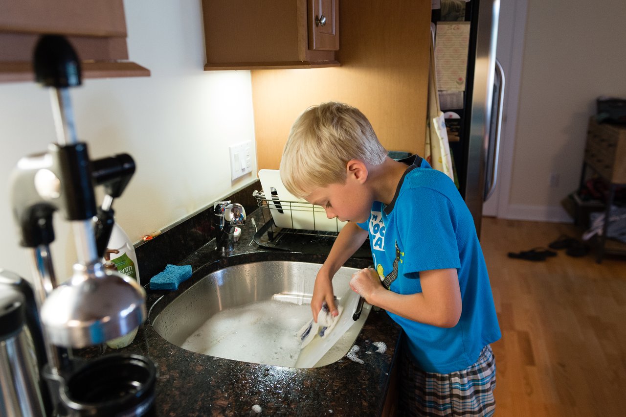 A child in a blue shirt washes dishes in the kitchen sink with soapy water.