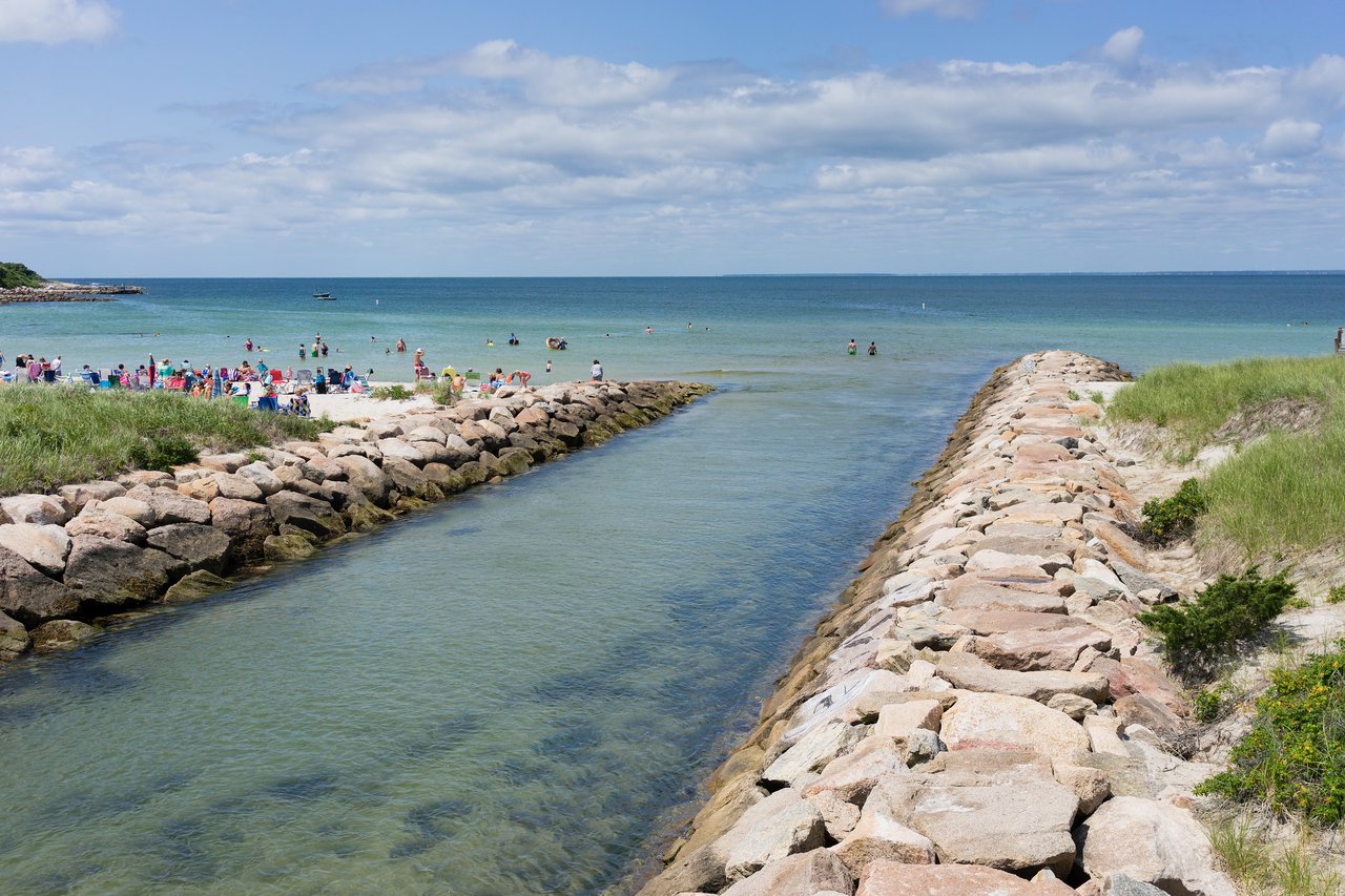 A rocky jetty divides a shallow inlet and a crowded beach, where people are swimming and relaxing under a sunny sky.
