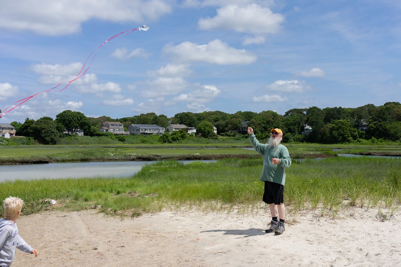 A man in a green shirt flies a kite with red tails while a child watches on a sandy beach.