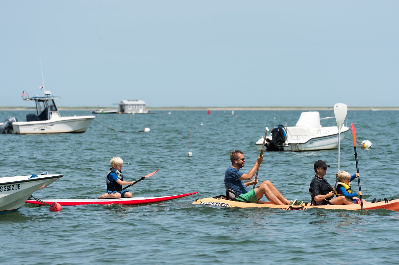 A group of people paddles on the water, with a child on a paddleboard and others in a kayak.