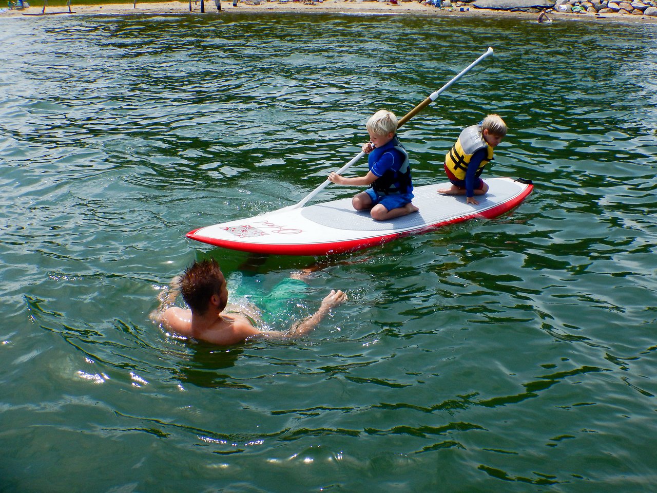 Two children wearing life jackets sit on a paddleboard, one holding a paddle, while an adult swims nearby.