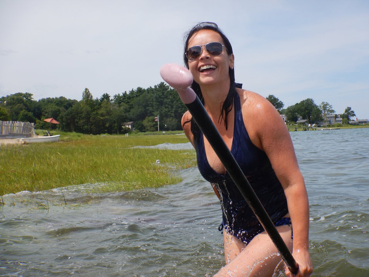 A smiling woman in a swimsuit holds a paddle while standing in shallow water on a sunny day.