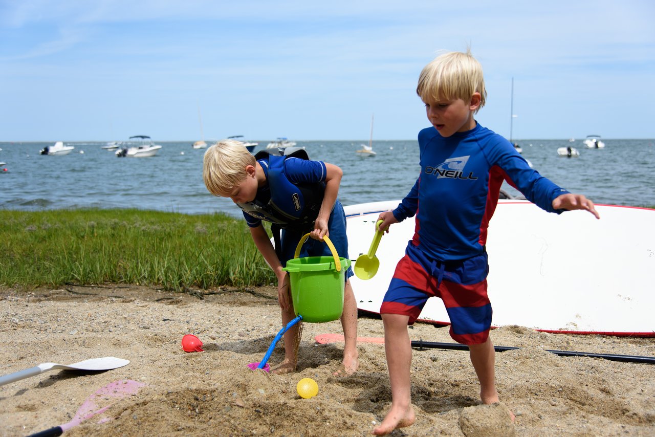 Two young children in swimwear play with sand toys on a beach, with a paddleboard and boats behind them.