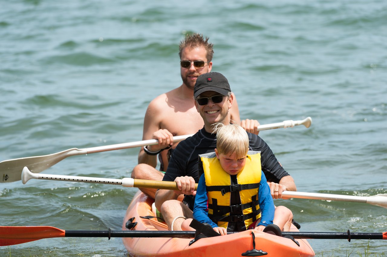 A child and two adults paddle a kayak on the water, with the child wearing a yellow life jacket.