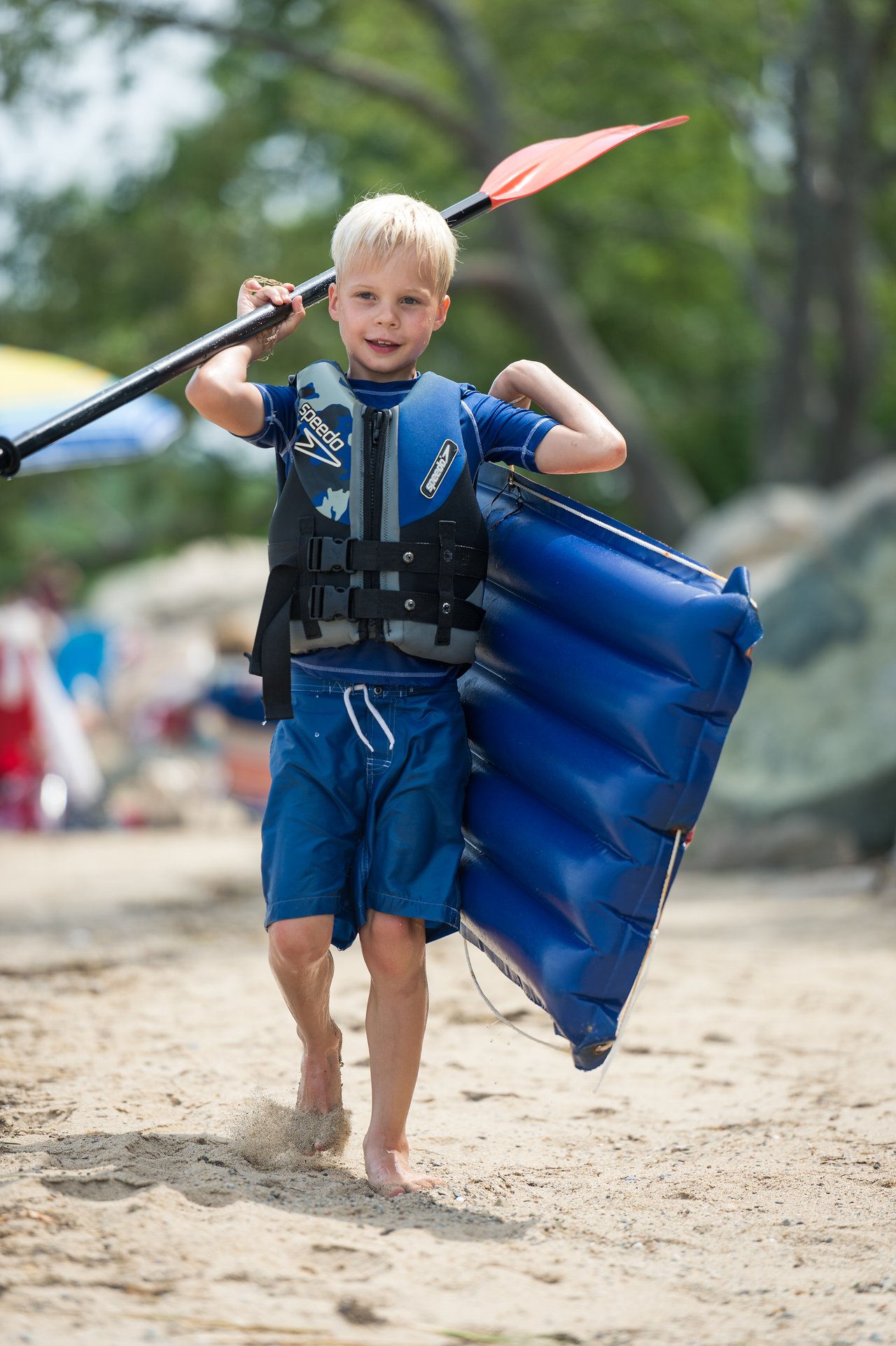 A young child in a life vest walks on the sandy beach, carrying a paddle and an inflatable raft.