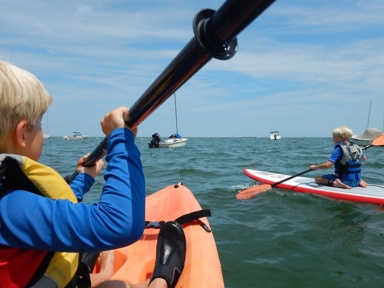 A child paddles a kayak while another child on a paddleboard moves through the water, both wearing life jackets.