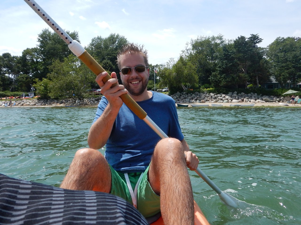 A man in sunglasses and a blue shirt paddles a kayak on the water, smiling at the camera.