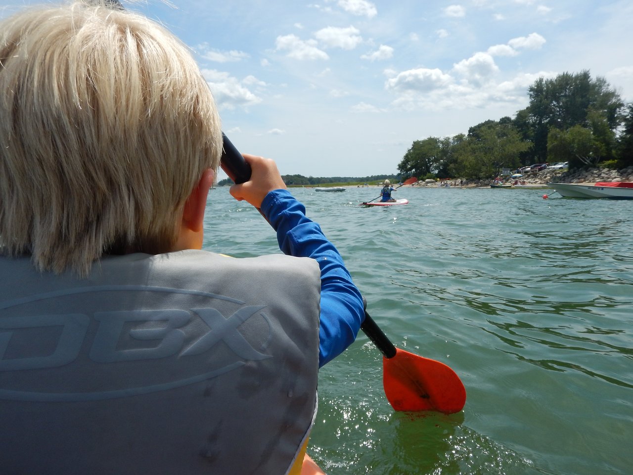 A person wearing a life jacket paddles a kayak on the water, with another kayaker visible in the distance.