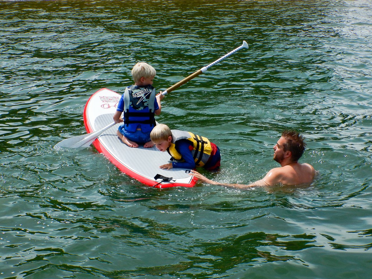 Two children wearing life jackets sit on a paddleboard while an adult swims nearby in the water.