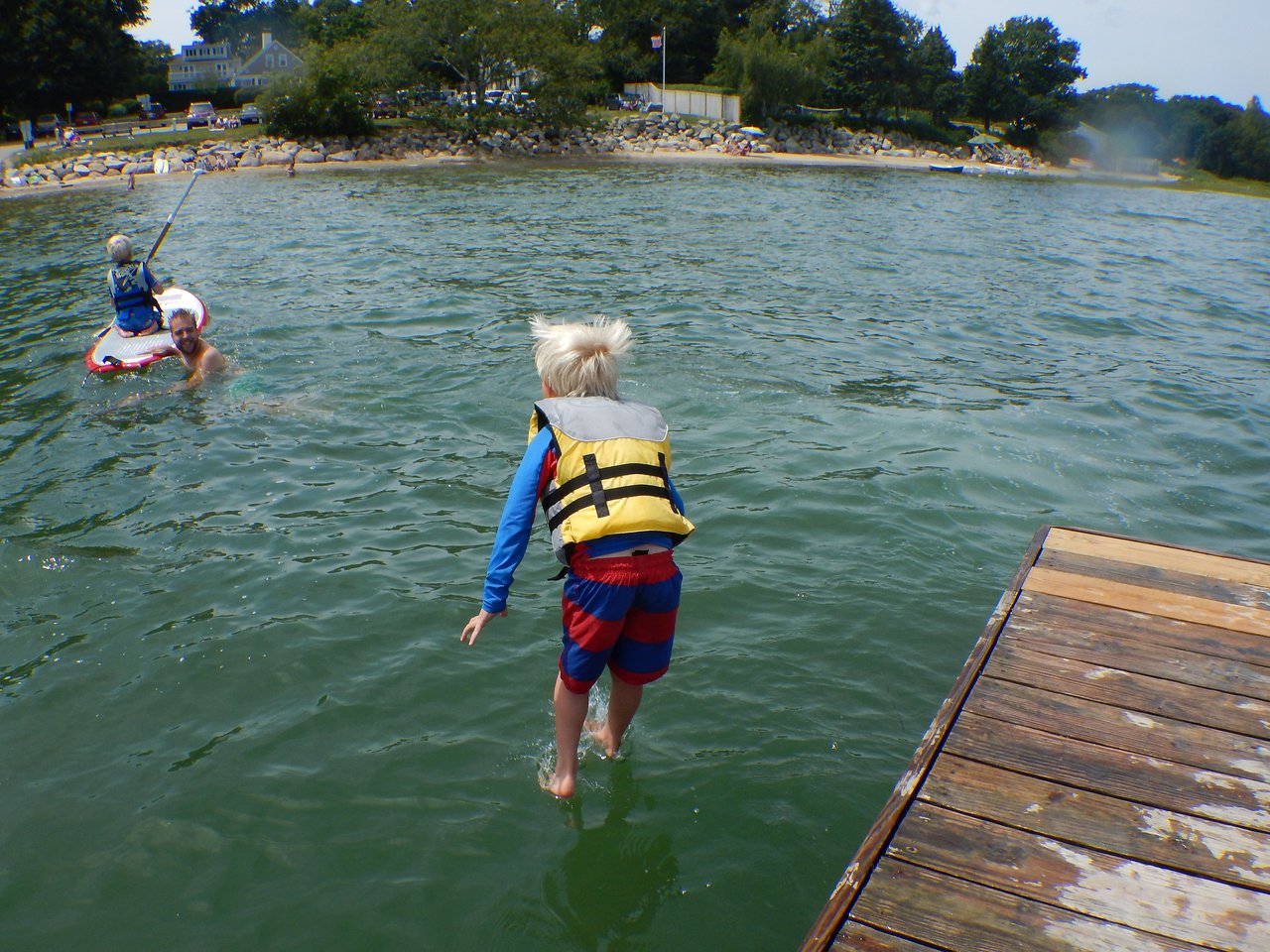 A child in a life jacket jumps off a dock into the water while others paddle nearby.