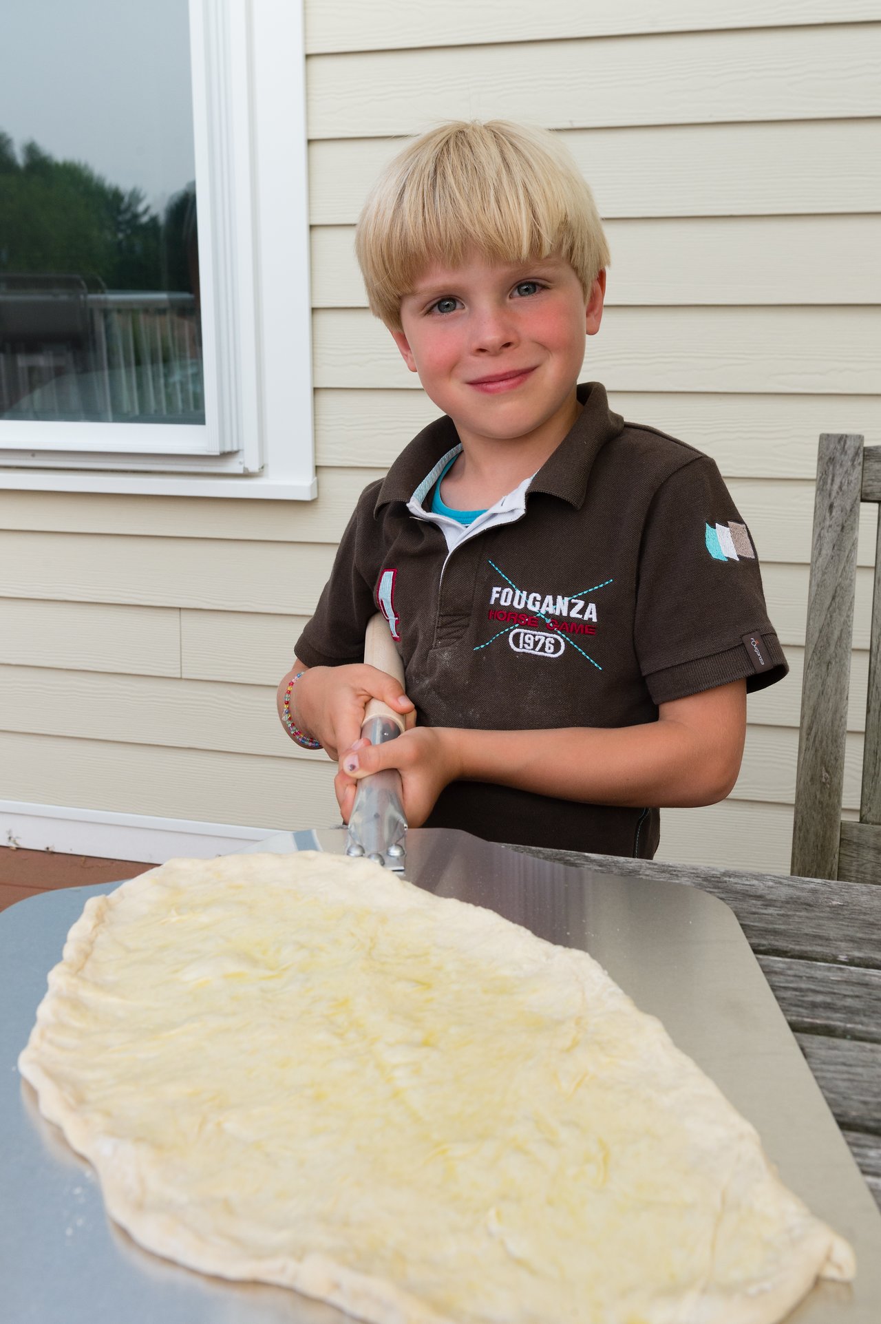 A child holds a pizza cutter over uncooked pizza dough on a metal tray, preparing to grill.