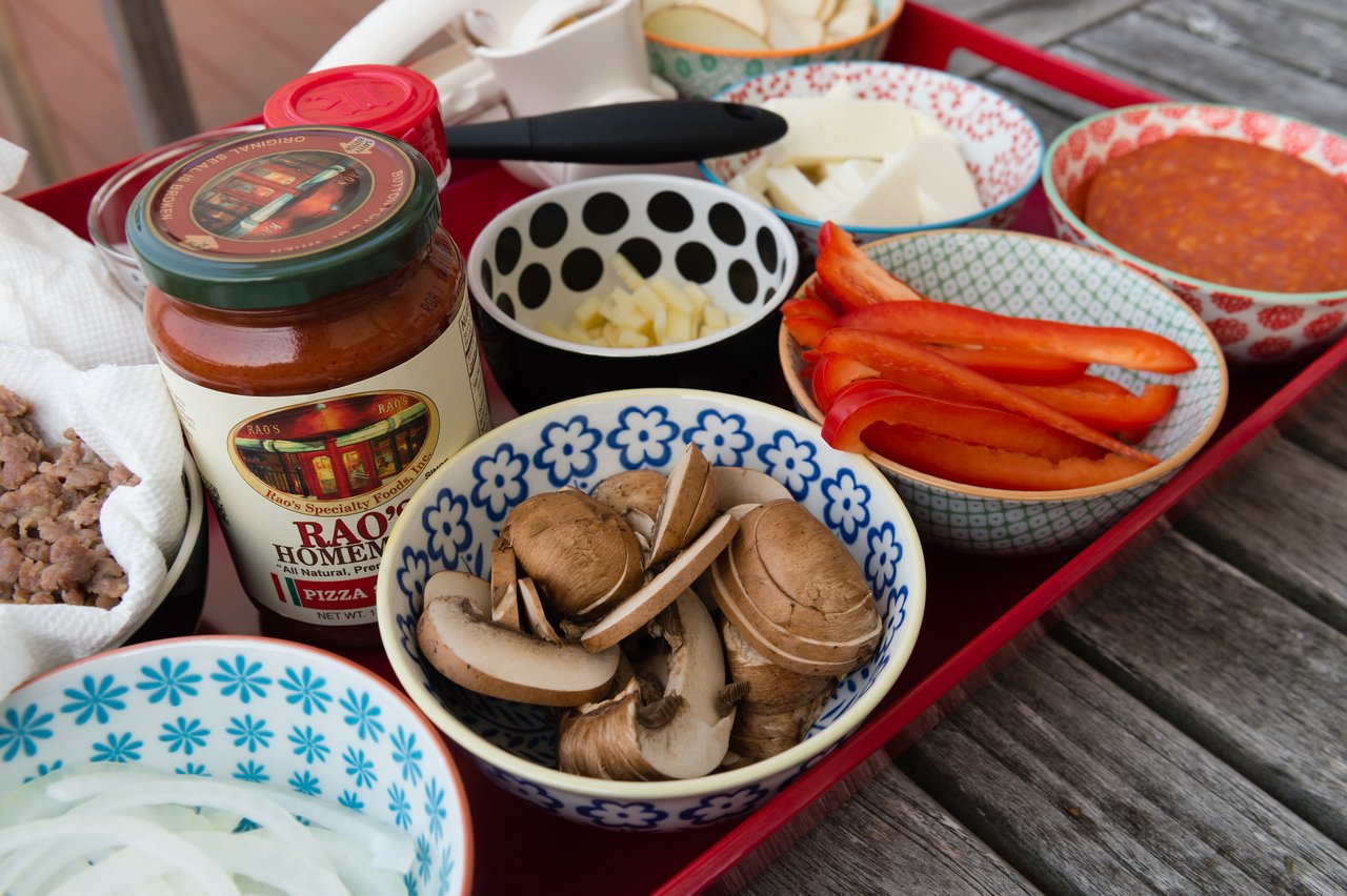 A tray with bowls of pizza toppings, including mushrooms, red peppers, onions, cheese, sausage, and a jar of sauce.