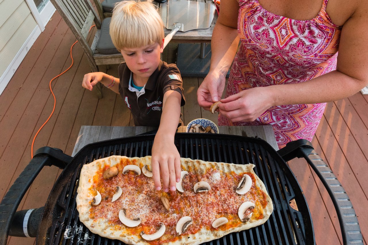 A child and an adult add mushrooms to a homemade pizza cooking on a grill outdoors.