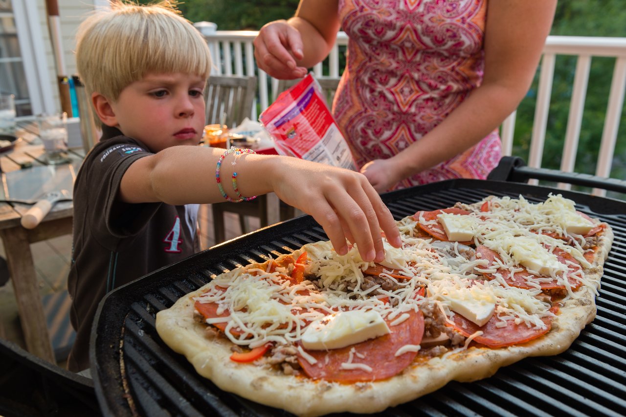 A child adds cheese to a homemade pizza cooking on a grill, with an adult assisting nearby.