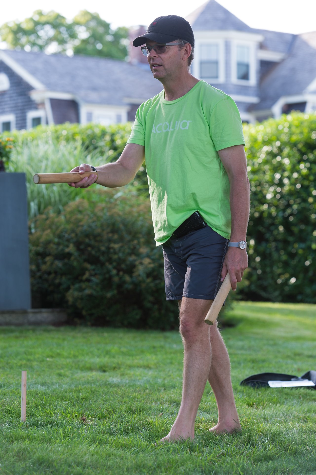 A man in a green shirt plays kubb, preparing to throw a wooden baton on a grassy lawn.