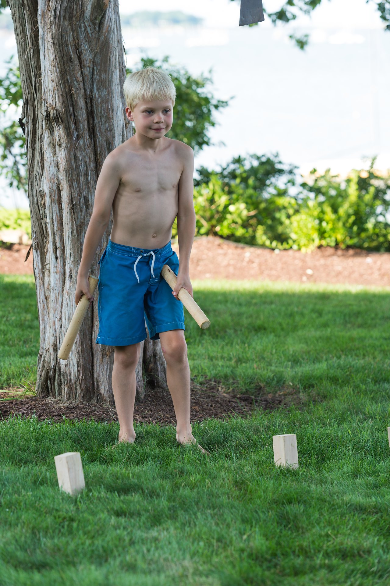 A boy in blue shorts holds wooden batons while playing kubb on a grassy lawn.