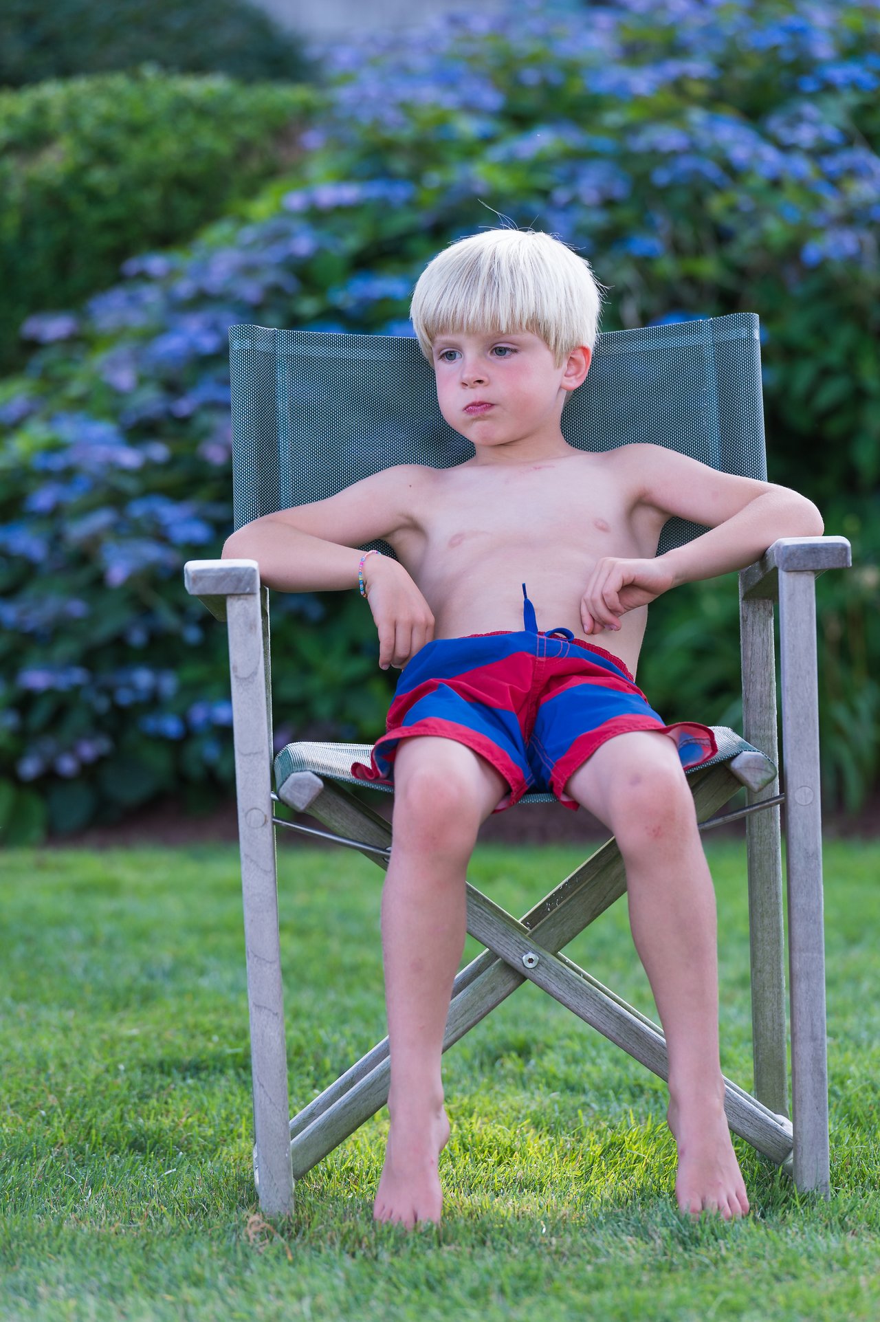 A young boy sits on a folding chair in the grass, watching a game of Kubb.