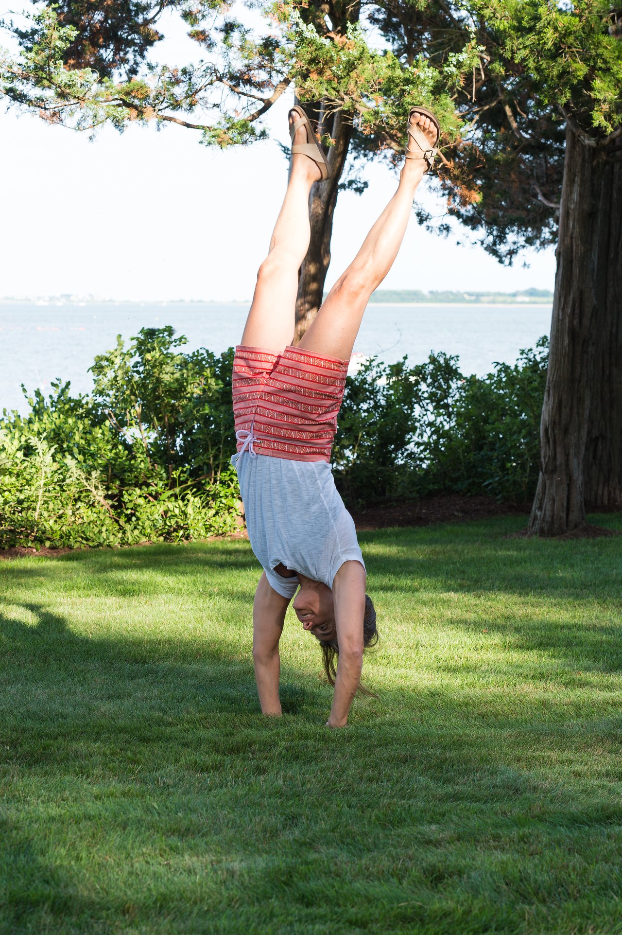 A person does a handstand on the grass near trees and water, wearing a gray shirt and red shorts.