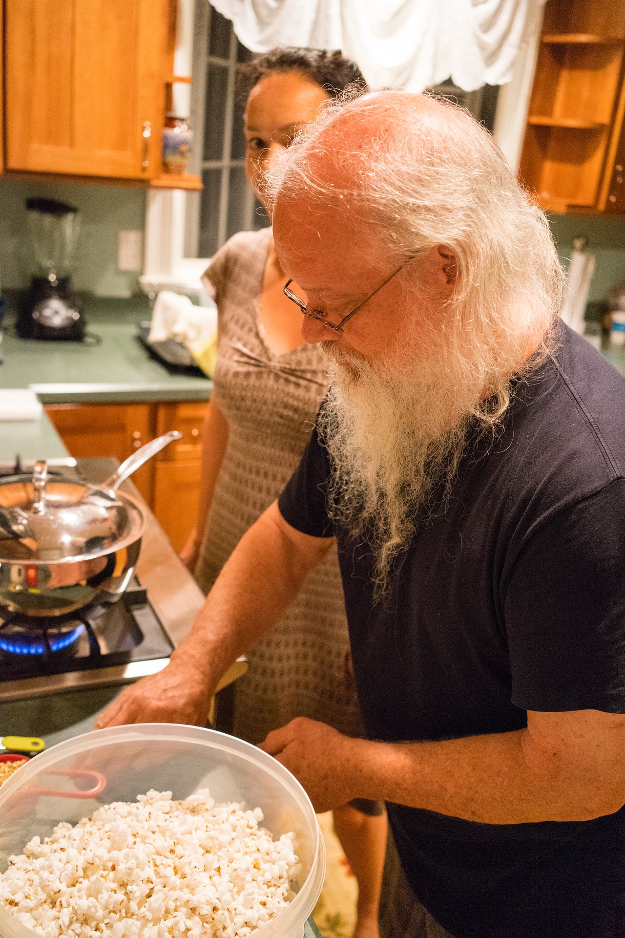 A man prepares freshly popped popcorn in a kitchen, with a pan on the stove and a woman nearby.