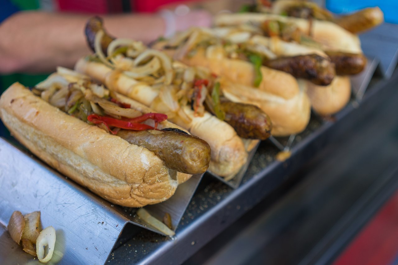 Several grilled sausages in buns topped with sautéed onions and peppers, served on a metal tray.
