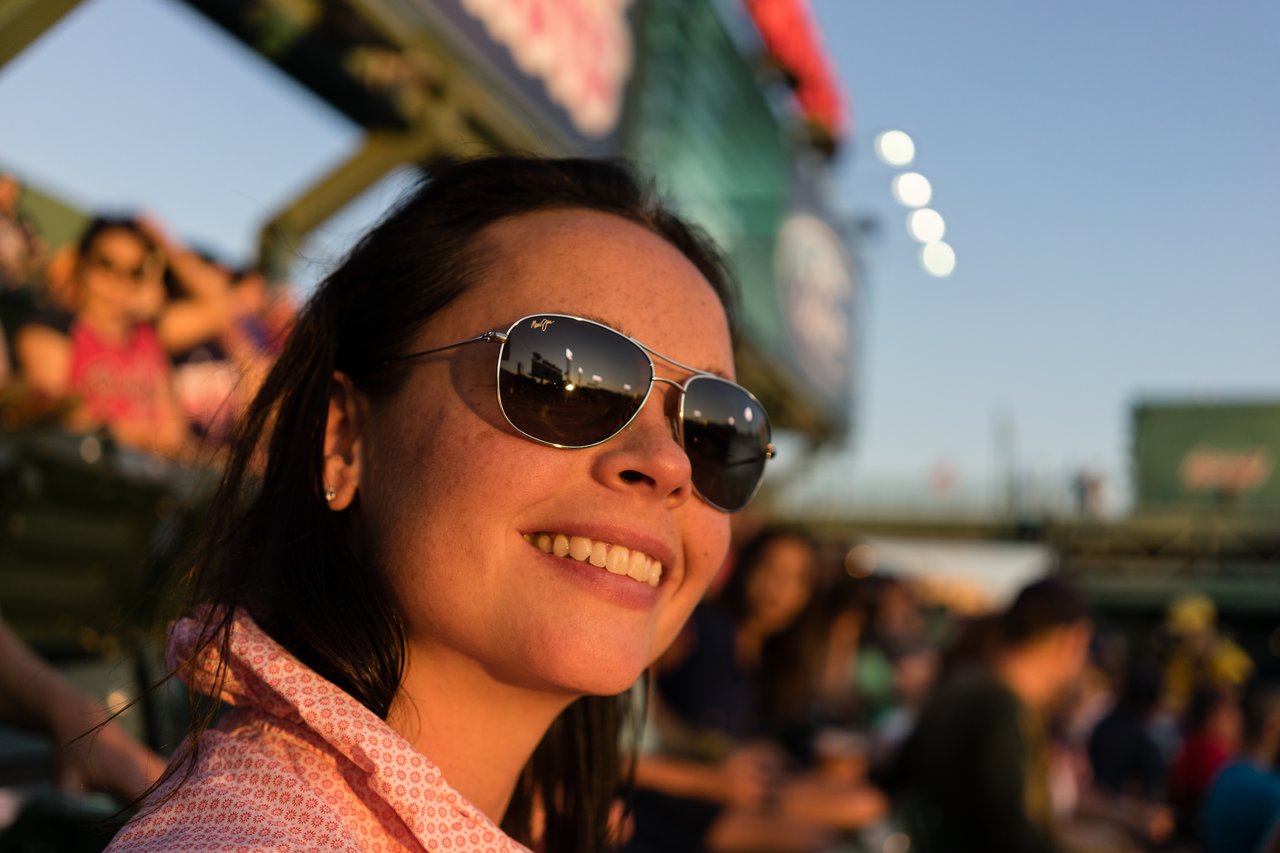 A woman wearing sunglasses smiles at a baseball stadium, with the game reflected in her lenses.