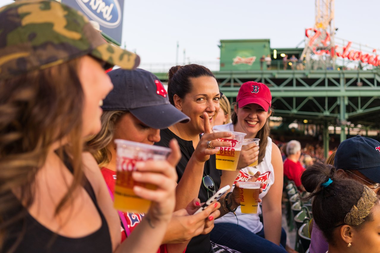 A group of people in Red Sox gear enjoying drinks and smiling at a baseball game.