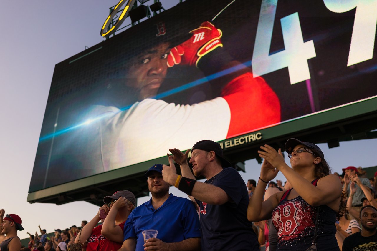 Red Sox fans cheer in the stadium, with a large screen showing a baseball player preparing to swing.