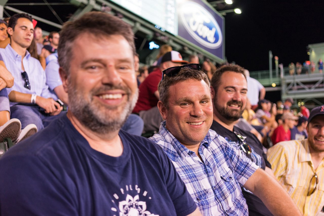 A group of smiling men sitting in the stands at a Red Sox game at night.