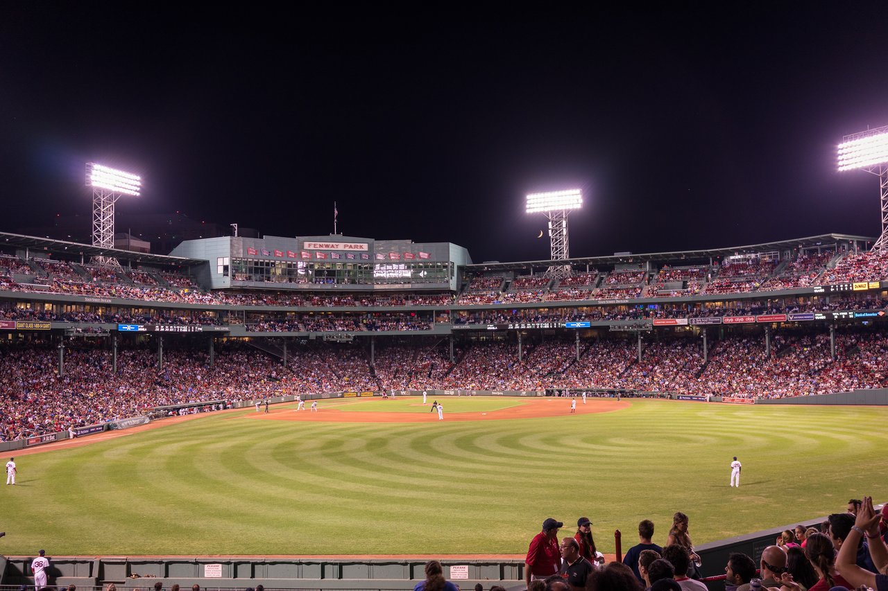 A nighttime baseball game at Fenway Park with players on the field and a crowd filling the stands.