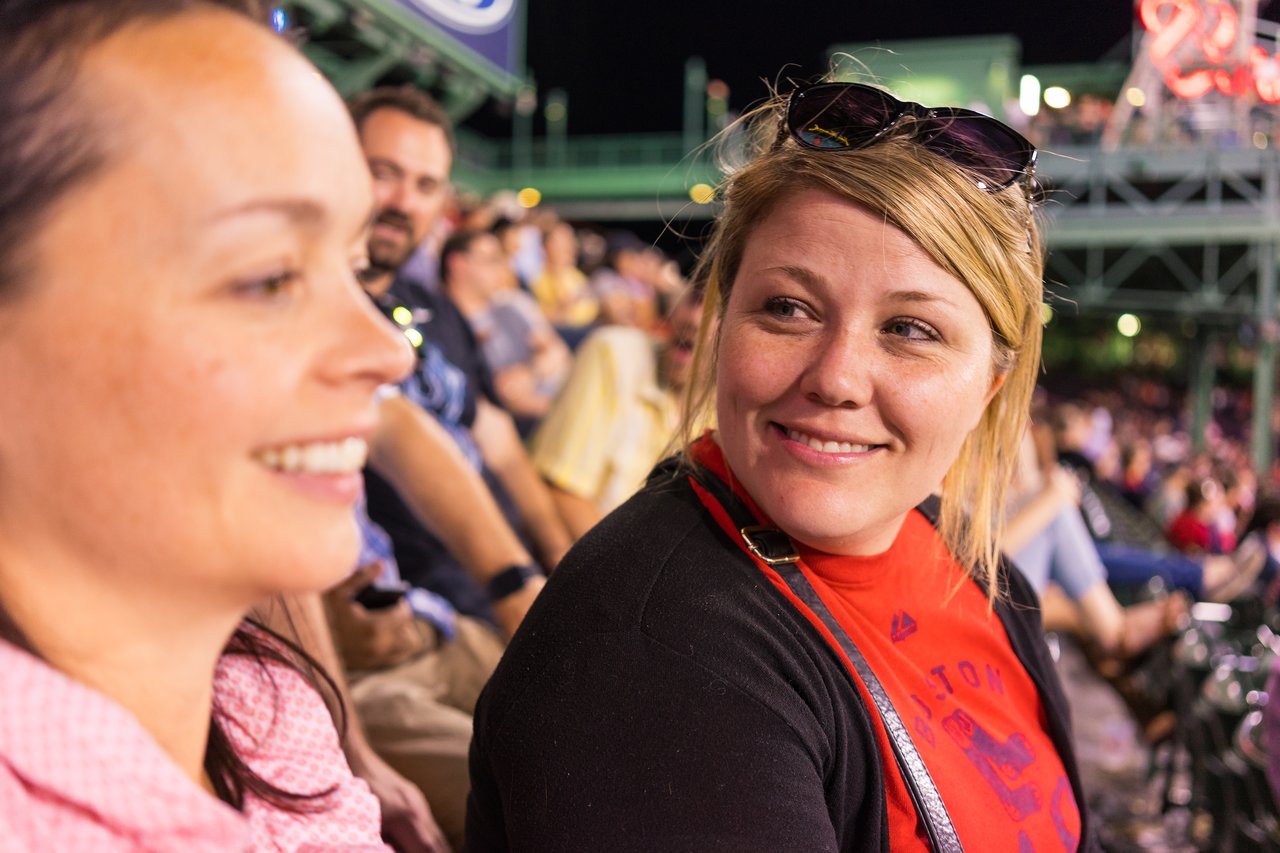 Two women sitting in a crowded stadium, one smiling and wearing a Boston Red Sox shirt.