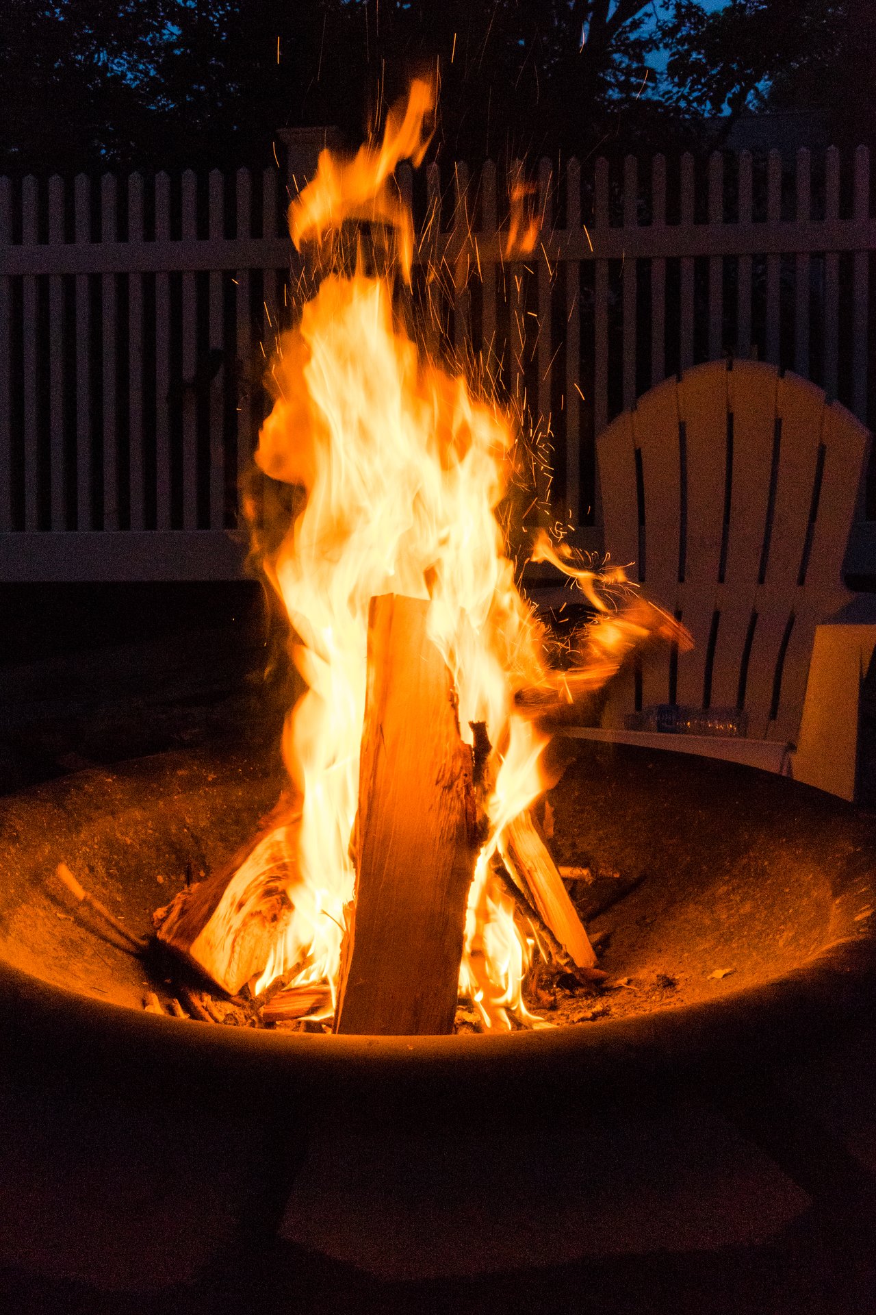 A campfire burns in a fire pit at night, with chairs nearby for gathering.